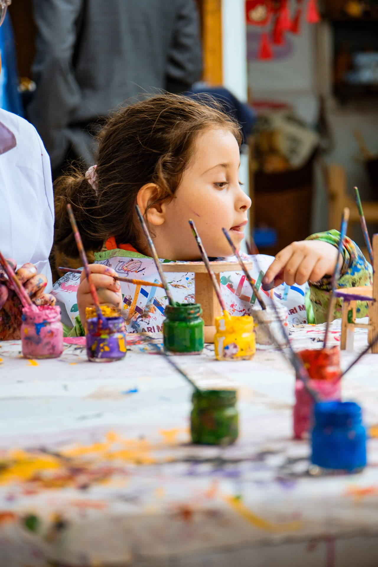 A girl painting at a busy art class table filled with paint jars and brushes