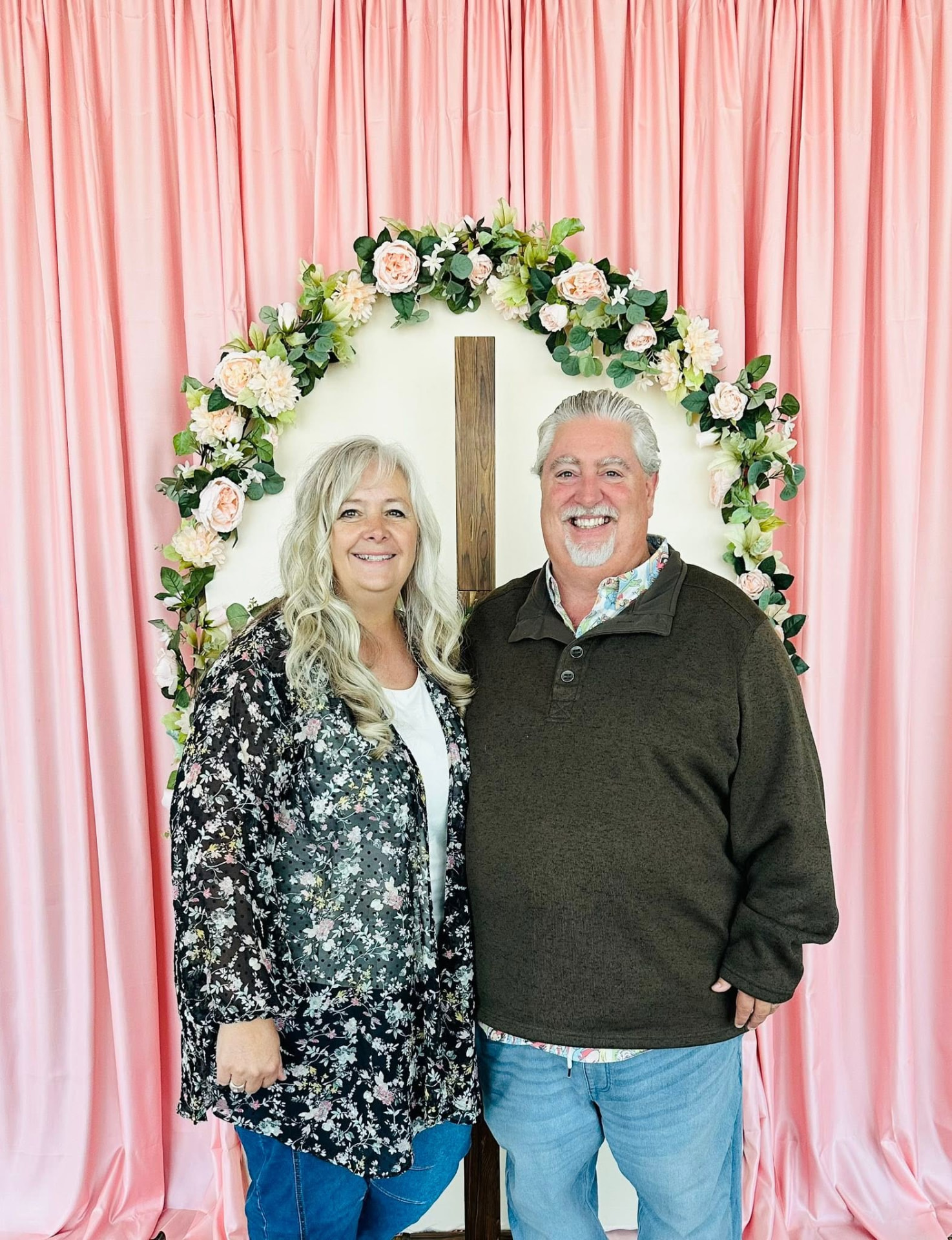 Jeff and Paula, founders of Afterschool America, standing together in front of a floral arch and wooden cross